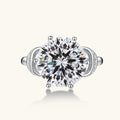 Sterling silver ring with 5ct round moissanite center stone and intricate bow design with pavé accents on white background.