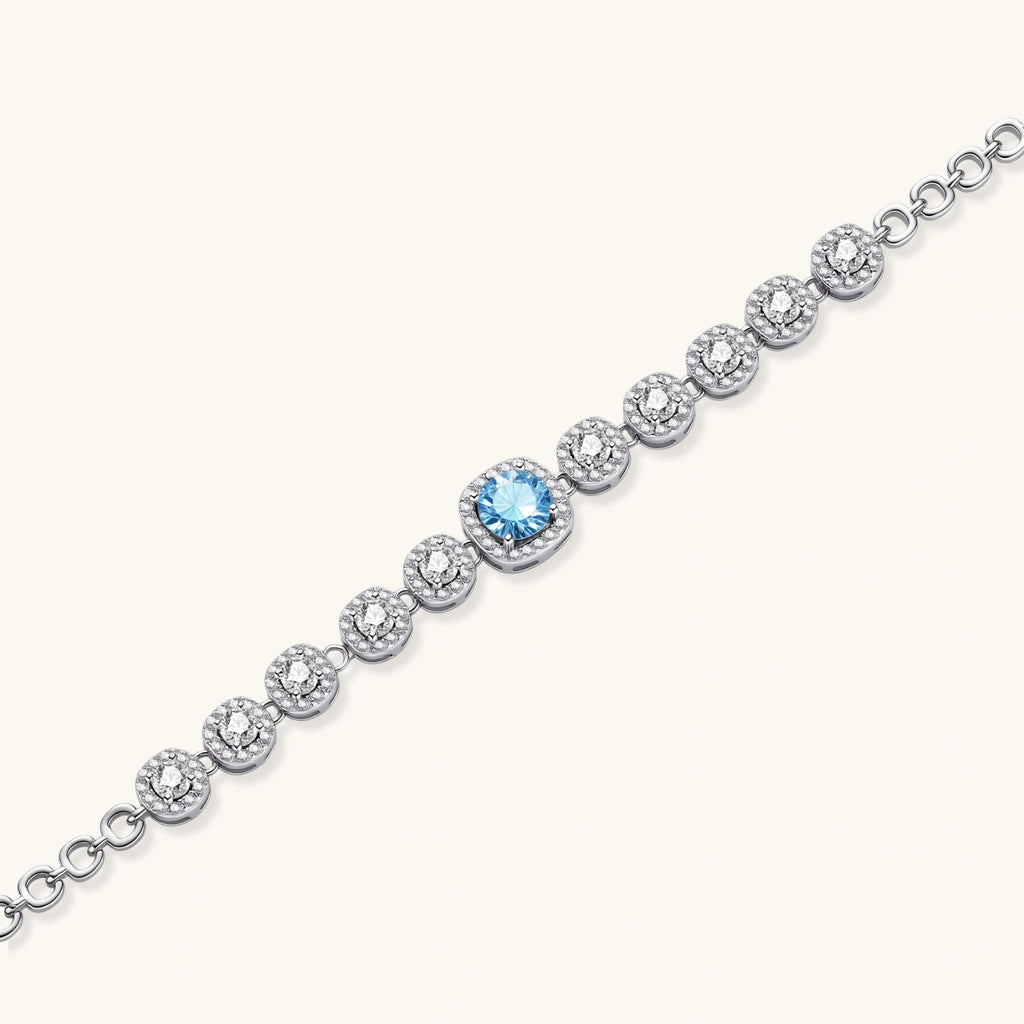 Sterling silver tennis bracelet with round halo-set moissanite stones and blue center stone on cream background.