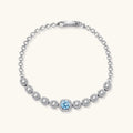 Sterling silver tennis bracelet with round halo-set moissanite stones and blue center stone on cream background.